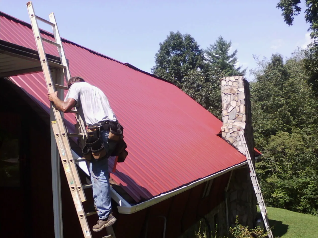 Roofer on a ladder beside a red metal roof and stone chimney — Soffit & Fascia Repair by Roofing Brothers in Milledgeville